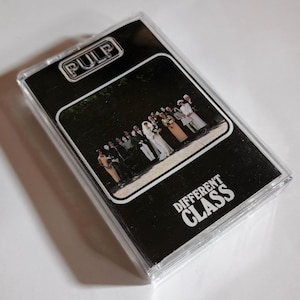 May include: A clear plastic cassette case containing the album Different Class by the band Pulp. The cover features a black background with the word PULP in a white outlined box at the top and the text DIFFERENT CLASS in white at the bottom.