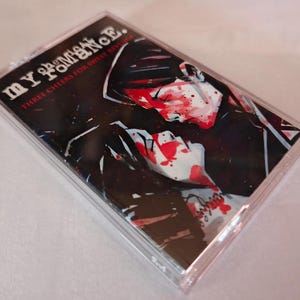 May include: A cassette tape in a clear plastic case. The cover art features the band name "MY CHEMICAL ROMANCE" and the album title "THREE CHEERS FOR SWEET REVENGE". The artwork depicts a dark, stylized image with red and white accents.