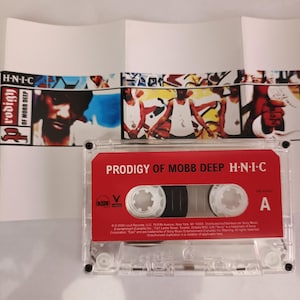 May include: A clear cassette tape with a red label bearing the text "PRODIGY OF MOBB DEEP H-N-I-C." The cassette is in front of a folded album cover with images of people and text. The album cover is white.
