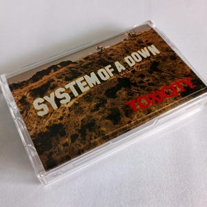 May include: A clear cassette tape case featuring the album cover for System of a Down's "Toxicity." The cover art depicts a brown, textured landscape with the band's name in white and the album title in red.