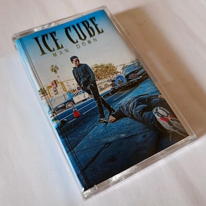 Puede incluir: Cinta de cassette vintage con el álbum "Man Down" de Ice Cube. La portada muestra una escena callejera con una persona de pie y otra en el suelo. El título y el nombre del artista están en blanco sobre un fondo azul.