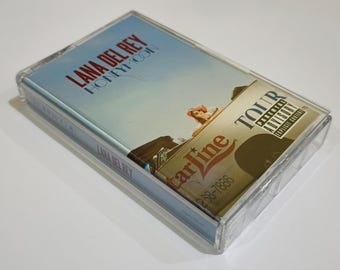 Puede incluir: Una cinta de casete del álbum "Honeymoon" de Lana Del Rey en una funda de plástico transparente. La carátula del álbum presenta un cielo azul claro y una mujer en un coche. La funda del casete también tiene una pegatina de "Tour".