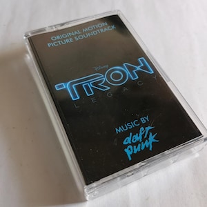 May include: A cassette tape of the "TRON: Legacy" original motion picture soundtrack. The black cassette case features the film's logo in blue neon lettering, with "MUSIC BY daft punk" in a matching font.