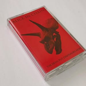 May include: A cassette tape with a red cover featuring a brown dinosaur skull graphic. The text "ALICE IN CHAINS" is at the top, and "THE DEVIL PUT DINOSAURS HERE" is at the bottom. The cassette is in a clear plastic case.