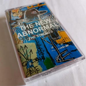 Könnte beinhalten: Eine Musikkassette mit einem farbenfrohen Cover, das den Text "THE NEW ABNORMAL" und "THE STROKES" zeigt. Das Artwork beinhaltet abstrakte Designs in Blau, Gelb und Weiß. Die Kassette befindet sich in einer transparenten Kunststoffhülle.