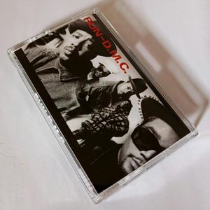 Puede incluir: Un casete vintage del grupo Run-DMC. La portada del álbum es en blanco y negro, con los miembros de la banda en varias poses. El texto "RUN-D.M.C." está en rojo, y "BACK FROM HELL" es visible.