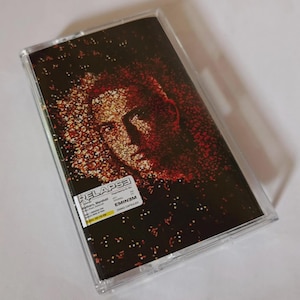 Puede incluir: Una cinta de cassette transparente con la portada del álbum "Relapse" de Eminem. La portada muestra un retrato de Eminem en tonos rojos y marrones, con el título del álbum y el nombre del artista.