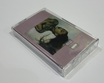 May include: A cassette tape with a clear plastic case. The cover art features a woman with long dark hair and a tattoo on her neck. The background is a muted pink color. The cassette has an advisory sticker.