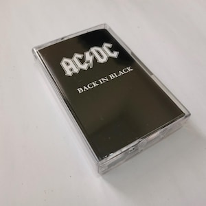 Pode incluir: Uma fita cassete preta com o texto "AC/DC" em branco acima de "BACK IN BLACK". A cassete está numa caixa de plástico transparente. A imagem é tirada numa superfície branca.