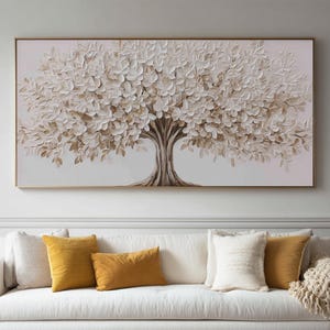 Pode incluir: Uma peça de arte de parede retangular com uma pintura texturizada de estilo impasto de uma árvore com folhas brancas e beges e um tronco castanho, emoldurada por uma borda dourada fina, exibida sobre um sofá branco com almofadas amarelas e brancas.