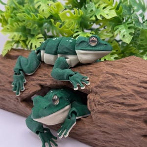 May include: Two articulated green and white frog figurines with realistic eyes, positioned on a textured brown log. The top frog is perched on the surface, while the second frog is partially visible within a hollow opening in the log.