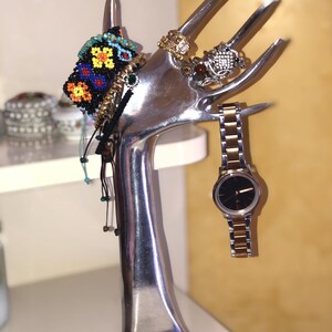 May include: A silver-colored hand-shaped jewelry holder displaying various accessories. It holds a beaded bracelet, rings, and a watch with a black face and gold and silver band. The background is a light yellow.