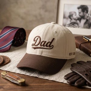Puede incluir: Gorra de béisbol beige y marrón con la palabra "Dad" bordada en la parte delantera. La gorra tiene una visera curva y el texto "EST. 20" en el lateral. Una corbata a rayas burdeos y azules es visible en el fondo.