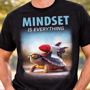 Può includere: T-shirt nero con la scritta "MINDSET IS EVERYTHING" in blu. La maglietta presenta una grafica di una tartaruga con occhiali da sole e un razzo sulla schiena. La persona nell'immagine indossa occhiali da sole e jeans blu.