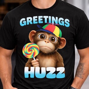 Op de afbeelding: Zwart T-shirt met de woorden "GREETINGS HUZZ" in blauw en wit. Het shirt heeft een cartoon aap met een kleurrijke hoed en een regenboog lolly. De aap heeft grote bruine ogen.