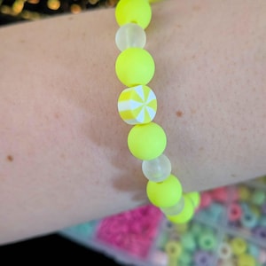 Pode incluir: Uma pulseira de contas amarelo néon com uma mistura de contas redondas e estampadas. A pulseira apresenta uma única conta estampada com um design de estrela branca e amarela. A pulseira é feita de contas redondas e lisas.