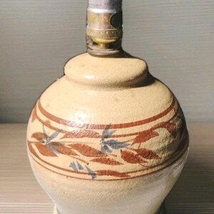 Pode incluir: Uma base de lâmpada de cerâmica bege com corpo redondo e pescoço estreito. A base é decorada com desenhos de folhas e galhos marrons e azuis. A lâmpada tem um encaixe de metal na parte superior.