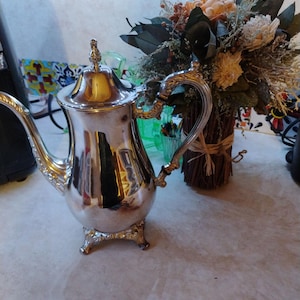May include: A silver-colored teapot with ornate detailing, including a curved spout and decorative handle. The teapot sits on small, decorative feet. A bouquet of dried flowers is in the background.