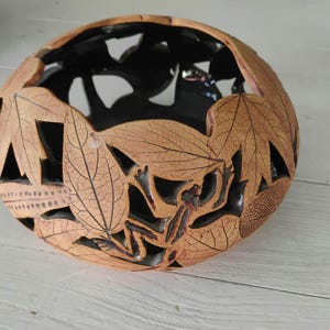 May include: A round, hollowed-out bowl with a tan, textured exterior featuring intricate cutouts of leaves and a frog. The interior of the bowl is finished in a smooth, glossy black glaze, creating a contrast with the natural-toned outer surface.