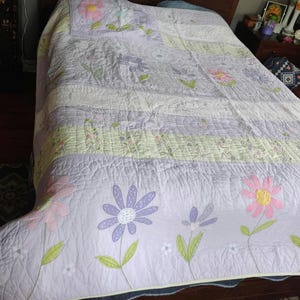 May include: A floral quilt in shades of lavender, white, and green, featuring large embroidered flowers. The quilt is spread over a bed, showcasing its patchwork design and decorative edge. The flowers are in pink, purple, and yellow.