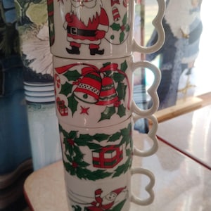 Könnte beinhalten: Ein Stapel von vier weißen Weihnachtsbechern aus Keramik. Jeder Becher zeigt ein anderes festliches Design, darunter der Weihnachtsmann, Glocken, ein Geschenk und der Weihnachtsmann im Schlitten. Die Becher haben herzförmige Griffe und sind mit roten, grünen und weißen Akzenten verziert.