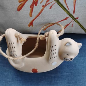 May include: A beige ceramic hanging planter shaped like a cat with a smiling face, small dark markings on its limbs, and a single orange dot on its side. The planter features a hollow center and is suspended by a natural jute twine rope.