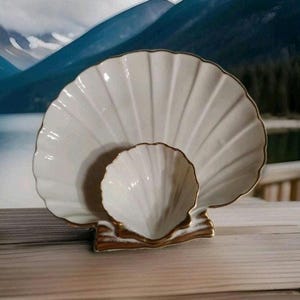 Può includere: Servizio di piatti a forma di conchiglia in ceramica color crema con finiture dorate. Il set comprende un piatto grande a forma di ventaglio e una ciotola più piccola abbinata. La base ha una finitura marrone strutturata.