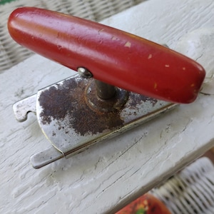 May include: A vintage can opener featuring a red wooden handle and a silver metal body. The handle shows signs of wear, with some paint chipping. The metal has some rust. The can opener is designed to open cans.
