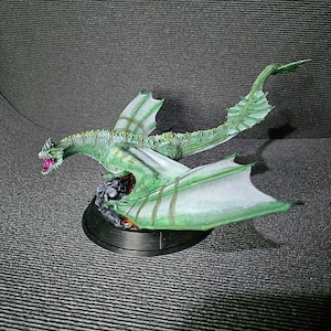 May include: A green dragon figurine with a textured, scaly body, large pale green wings with visible structural veins, and an open mouth revealing a bright pink interior. The creature is mounted on a circular black base atop dark rocks.