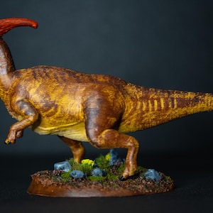 Pode incluir: Figura detalhada de um dinossauro Parasaurolophus. O modelo apresenta um corpo castanho e amarelo com uma crista vermelha distinta. Está sobre uma pequena base texturizada com rochas e vegetação em miniatura, mostrando uma cena pré-histórica realista.