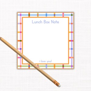 May include: A square lunch box note with a colorful plaid border in orange, pink, blue, and green. The words "Lunch Box Note" are at the top, and "I love you!" at the bottom. A wooden pencil rests diagonally across the note.