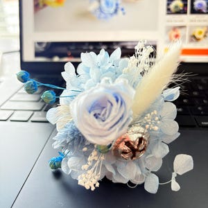 May include: A delicate light blue floral arrangement, featuring a central rose surrounded by blue petals and white and beige accents. A small, metallic bell adds detail. Likely for a wedding or special occasion, the arrangement is a charming accessory.