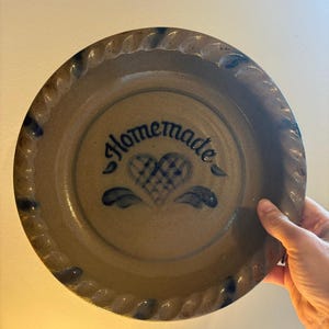 May include: A round, tan stoneware pie dish with a decorative rope-style rim. The center features the word "Homemade" in dark blue script, positioned above a blue heart graphic with a cross-hatch pattern and two small leaf-like accents.
