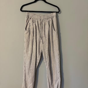 May include: A pair of striped pants hanging on a black hanger. The pants have a drawstring waist and two front pockets. The fabric is white with vertical gray stripes. The pants are made of a lightweight material.