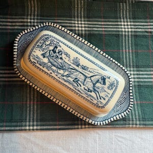 May include: A rectangular ceramic butter dish with a cream-colored base and a matching lid. The lid features a dark blue transferware illustration of a horse-drawn sleigh with two figures. The dish has a decorative scalloped edge with blue trim.