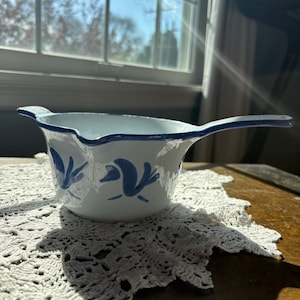 May include: White ceramic gravy boat with a blue rim and handle, decorated with blue floral designs. The gravy boat is placed on a white, crocheted doily, and sits on a wooden surface.