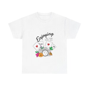 Puede incluir: Camiseta blanca con un diseño gráfico que presenta una cámara, naipes y flores. El texto "Enjoying my time with you" está escrito en una fuente de guion por encima del gráfico. El diseño incluye corazones y elementos florales.