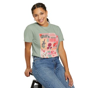 Puede incluir: Camiseta verde claro con un collage de diseños de sellos postales. Los sellos muestran ilustraciones florales, las palabras "LOVE CITY", "LOVE TO GALORE" y "XOXO". La camiseta se combina con vaqueros azules.