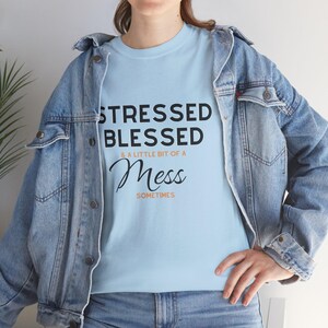 Puede incluir: Camiseta azul claro con las palabras "STRESSED BLESSED & A LITTLE BIT OF A Mess SOMETIMES" en letras negras. La camiseta se lleva con una chaqueta vaquera.