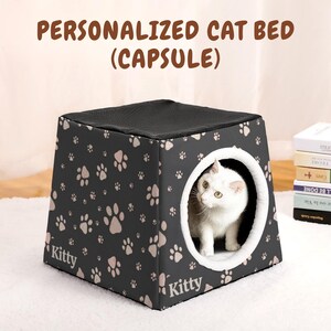 May include: A black, capsule-shaped cat bed with a paw print pattern and the word "Kitty". The bed has a dark gray mesh top and a white cat is visible inside the opening. The text "PERSONALIZED CAT BED (CAPSULE)" is at the top.