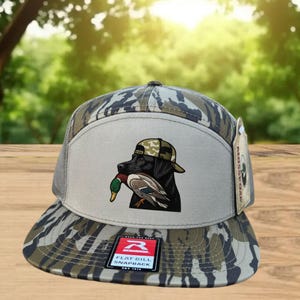 May include: A flat bill snapback hat with a camouflage pattern on the bill and crown. The hat features a graphic of a black Labrador wearing a camo hat and holding a duck. The hat has a mesh back and a Richardson tag.