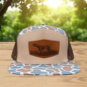 May include: A brown and tan trucker hat with a camouflage pattern on the bill. The hat features a leather patch with a dog illustration. The hat has a brown mesh back and a tan front panel.