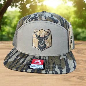 Może przedstawiać: Czapka typu snapback z płaskim daszkiem i wzorem kamuflażu na daszku i koronie. Przedni panel jest beżowy z emblematem głowy jelenia wewnątrz sześciokąta. Czapka ma siatkowy tył.