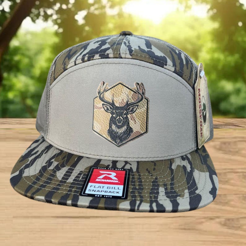 Może przedstawiać: Czapka typu snapback z płaskim daszkiem i wzorem kamuflażu na daszku i koronie. Przedni panel jest beżowy z emblematem głowy jelenia wewnątrz sześciokąta. Czapka ma siatkowy tył.
