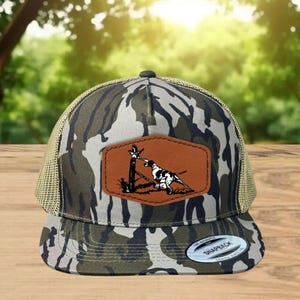 May include: Camouflage trucker hat with a brown leather patch featuring a black and white dog illustration. The hat has a mesh back and a curved brim. The hat is made of a camouflage pattern fabric.