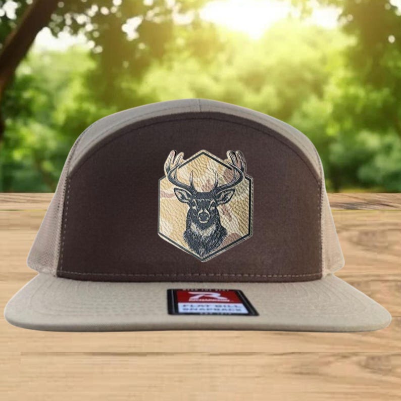 Może przedstawiać: Brązowo-beżowa czapka typu trucker z emblematem jelenia. Czapka ma brązowy panel przedni z sześciokątną naszywką przedstawiającą głowę jelenia. Siatkowa część tylna i daszek są beżowe. Czapka ma płaski daszek i metkę z napisem "Flat Bill Originals".