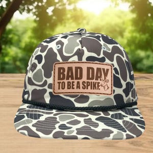 May include: A camouflage baseball cap with a brown leather patch. The patch reads "BAD DAY TO BE A SPIKE" in bold, capitalized letters. The cap features a black rope detail along the brim and a pattern of gray, white, and brown shapes.