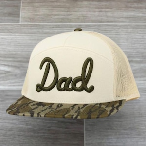 May include: Cream-colored baseball cap with the word "Dad" embroidered in dark green script. The brim features a camouflage pattern, and the back is a tan mesh.