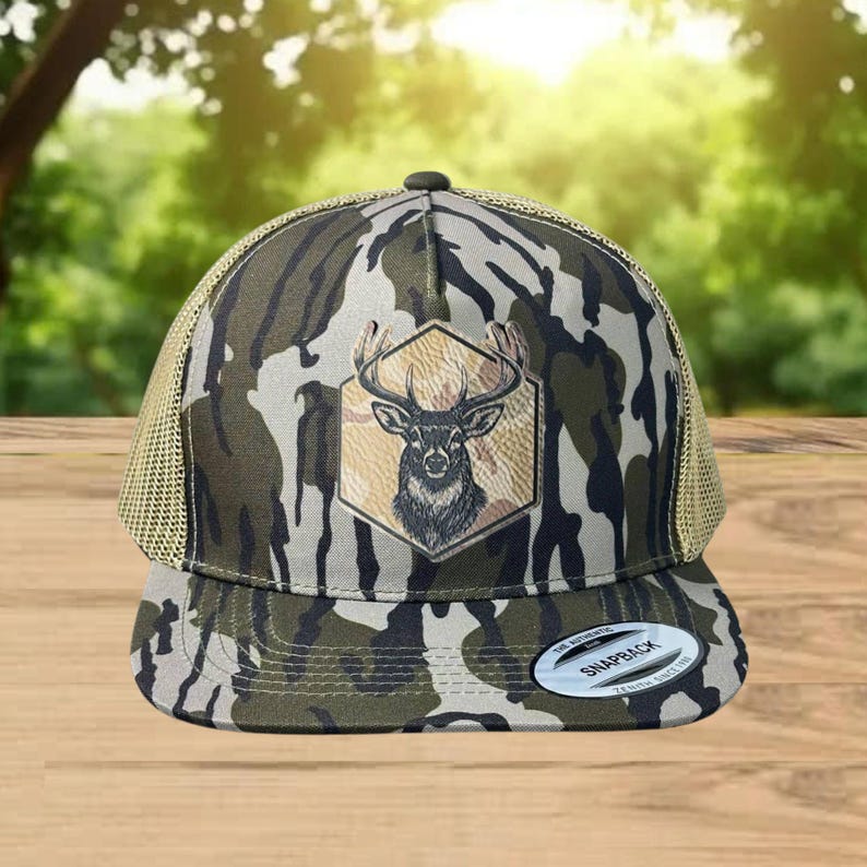 Może przedstawiać: Czapka typu snapback z wzorem kamuflażu i beżowym tyłem z siatki. Z przodu znajduje się brązowo-czarny emblemat głowy jelenia wewnątrz beżowego sześciokąta. Czapka ma zakrzywiony daszek i zapięcie na zatrzask.