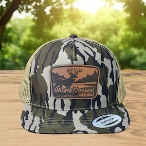 May include: A camouflage-patterned trucker hat with a brown leather patch featuring a deer silhouette and the text "Swamp Donkey." The hat has a mesh back and a curved brim. The hat is a snapback style.